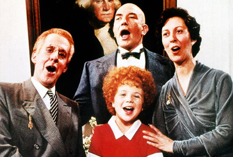 Scene from Annie, three adults gathered around a redheaded girl and singing together.