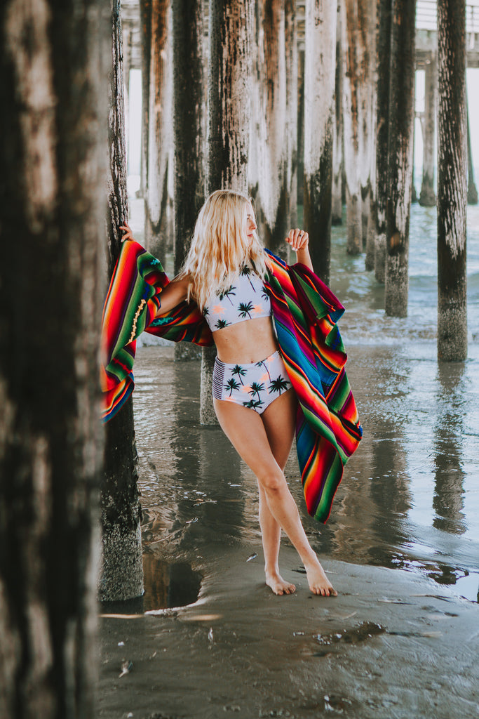 Alyssa walking under the boardwalk at the beach as she wears a palm-tree patterned bikini and a Hiptipico wide brim hat, wrapped in the Hiptipico Remolacha Blanket