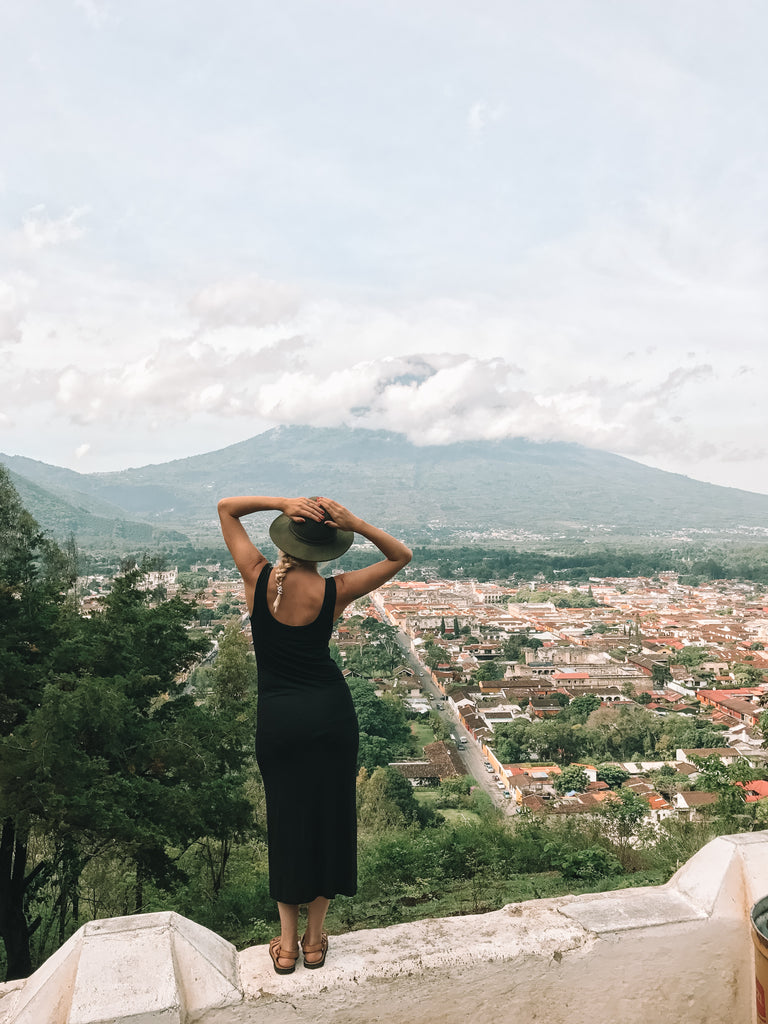Hiptipico founder Alyssa poses at lookout in black dress and olive hat in front of view of colonial Antigua cityscape and volcano, best views of Antigua, cityscape Antigua, top destinations Antigua Guatemala, must see Antigua Guatemala, ethical travel bloggers