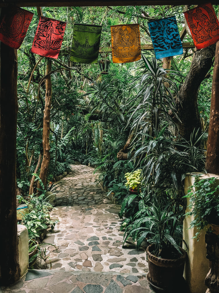 A brick pathway leads through lush wooded area with small cloth flags with spiritual figures at the top of the doorway, san marcos yoga, san marcos lake atitlan, san marcos sauna, where to do yoga by the lake, where to do yoga in Lake Atitlan, Lake Atitlan travel destinations, travel ethically in Lake Atitlan.