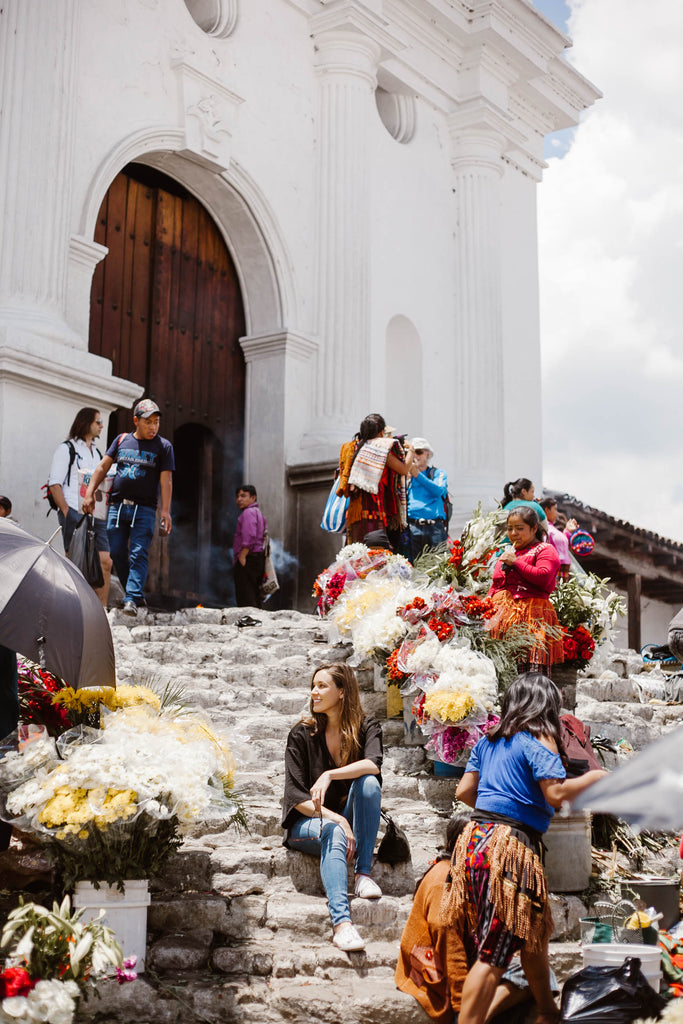 She Is Not Lost blogger Carina Otero sits smiling on Chichicastenango church steps surrounded by yellow, white and red bunches of flowers and vendors, Chichicastenango travel, Places to visit in Guatemala, must see guatemala travel, ethical travel in Guatemala, textile sourcing locations Central America