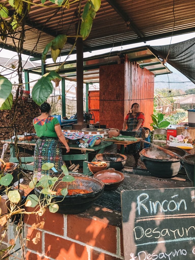 View of open air kitchen shows employees in traditional corte dresses, large bowls of traditional farm fresh foods, bowls with sauces, and lush vegetation, farm to table restaurants in Guatemala, top dining experiences in Antigua, most interesting places to visit Guatemala, ethical travel blogs