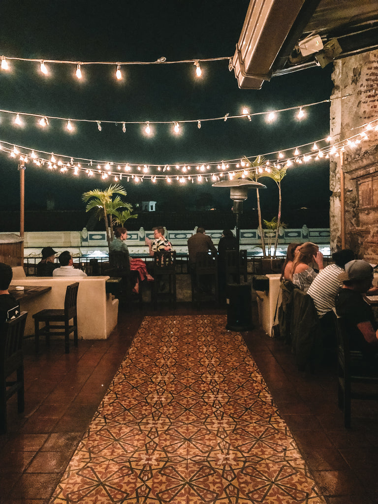People sit at roof top bar at Las Vibras with black chairs, beige walls, and detailed red brown geometric rug, party in Antigua, best places to part in Antigua, best night clubs in Antigua Guatemala, what should I do in Guatemala, ethical travel blogs, where to go for drinks Antigua