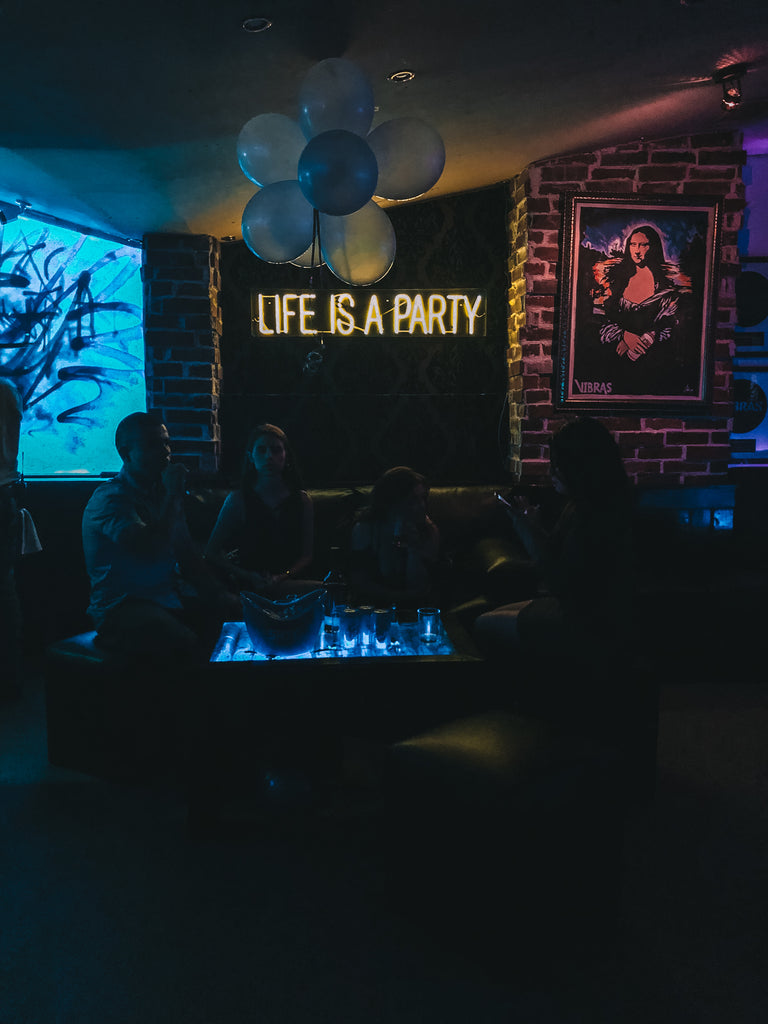 Customers sit on couches around lit coffee table storing drinks and ice under a neon sign that reads 'life is a party', coolest places to party in Guatemala, trendy nightclubs guatemala, trendiest spots Antigua, where should I go to party in Antigua