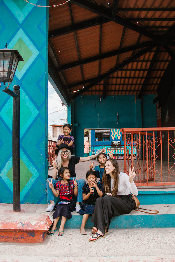 Hiptipico founder Alyssa and She Is Not Lost blogger Carina pose happily in blue and green painted town square with local children, authentic travel experiences in Guatemala, authentic travel Latin America, ethical travel options, ethical travel blogs