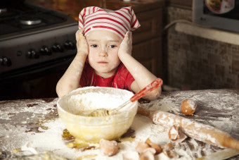 Little Girl wearing a baking hat with her hands on her face surrounded by flour and baking utensils