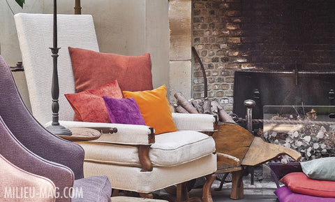 Raspberry colored cushions with violet and tangerine bring color and warmth to the antique armchair by the fire - Walda Pairon