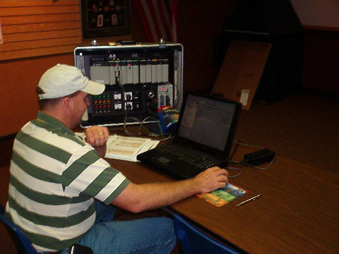 man in green and white striped shirt and white hat sitting at table with computer and electronics