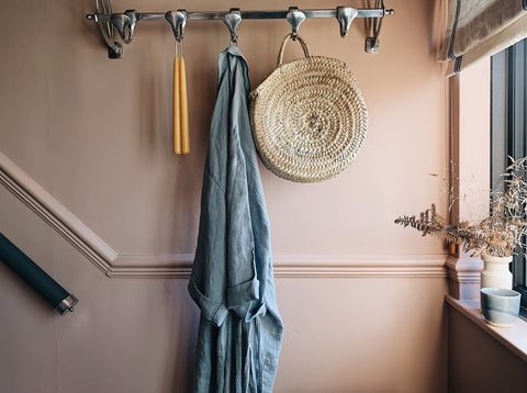 a pink wall with hooks on which a bag, denim dress and amber candles hang