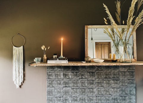 A mantlepiece is decorated with pampas grasses, candles and a woven wall decoration | Aerende