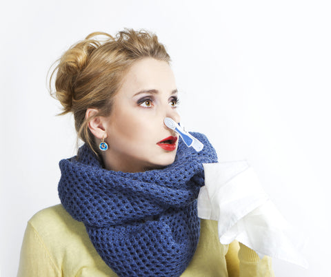 A woman in a blue scarf and yellow top looks off in the distance with a clothespin on her nose and a tissue in hand