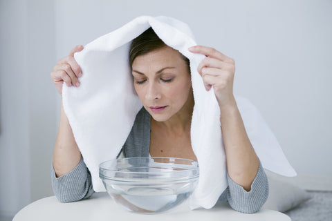 A woman holds a white towel over her head and inhales over a glass bowl