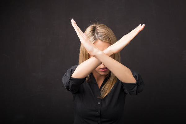 Woman standing in front of a black background wearing a black blouse crossing her arms like an 'X' in front of her face