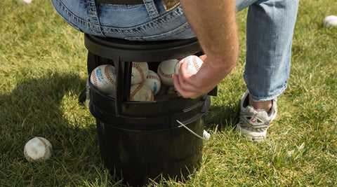 Baseball player sitting on The Original Bucket Stool™ on top of a bucket full of baseballs. Man reaching underneath to grab another ball for more efficient practice. Click to see baseball and softball solutions page.