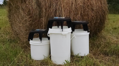 Three different sized buckets (3.5, 5, and 6-gallon buckets) with Bucket Stools™ on top to show how to adjust the height of the stool.  Hay bale on the farm in the background. Click to see farm and gardening solutions page.