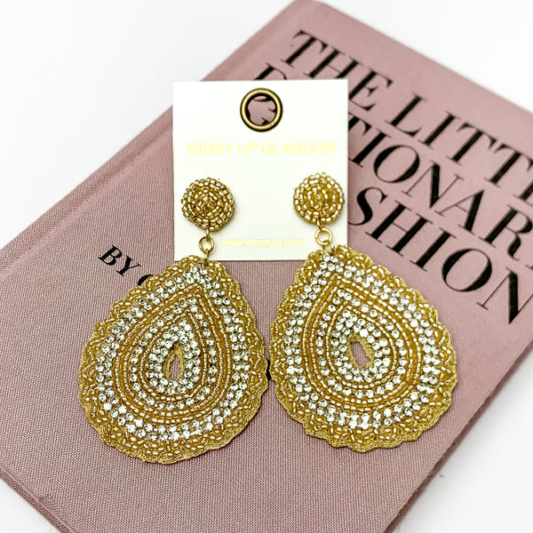 Sound Wave Beaded Drop Earrings with Clear Crystals in Gold. Pictured on a white background with a book behind the earrings.