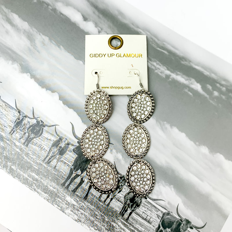 Three tier silver tone oval shape dangle earrings with clear crystals. Pictured on a black and white western picture.