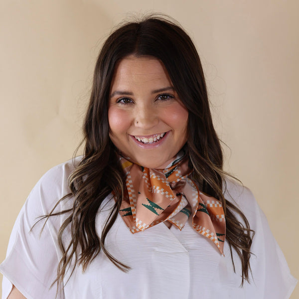 Brunette model wearing a white, drop shoulder top with tan scarf with a multicolor aztec design tied around her neck. Model is pictured in front of a beige background. 