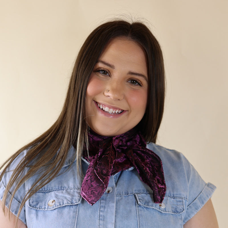 Brunette model is pictured wearing a denim button up top and a blackberry purple, baroque printed scarf tied around her neck. She is pictured in front of a beige background. 