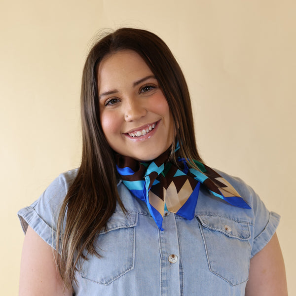 Brunette model wearing a blue dress with Blue and Brown print scarf tied around her neck. Model is pictured in front of a beige background.