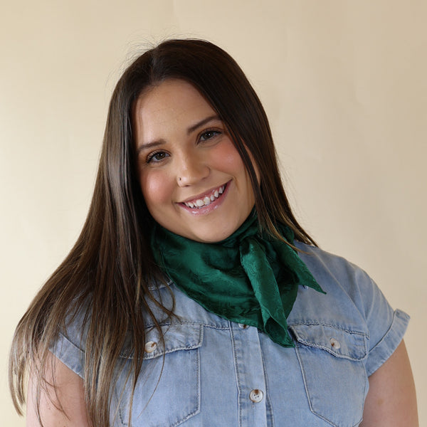 Brunette model wearing a short sleeve, denim button up with a forest green jacquard print scarf tied around her neck. This model is pictured in front of a beige background. 