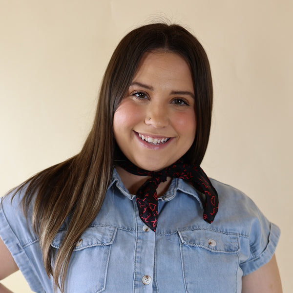 Brunette model is pictured wearing a denim button up top and a black scarf with a red print tied around her neck. She is pictured in front of a beige background. 