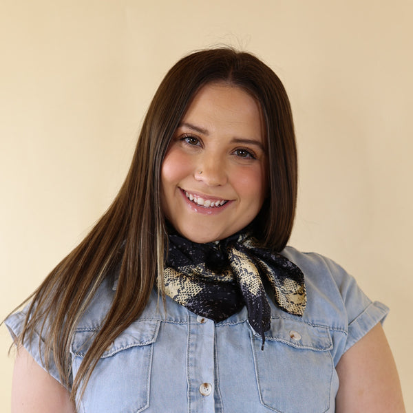 Brunette model wearing a short sleeve, denim button up with a black and white snake print scarf tied around her neck. This model is pictured in front of a beige background. 