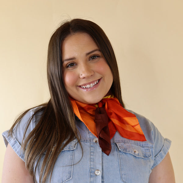 Brunette model wearing a blue dress with Ornage and Brown Mix Print scarf tied around her neck. Model is pictured in front of a beige background.