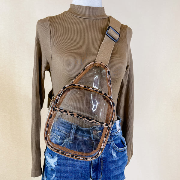 Pictured is a clear sling backpack with a leopard print outline. This bag also includes a tan strap and black and gold accents. This bag is on a mannequin that has blue denim shorts and a brown long sleeve shirt pictured in front of an ivory background.  