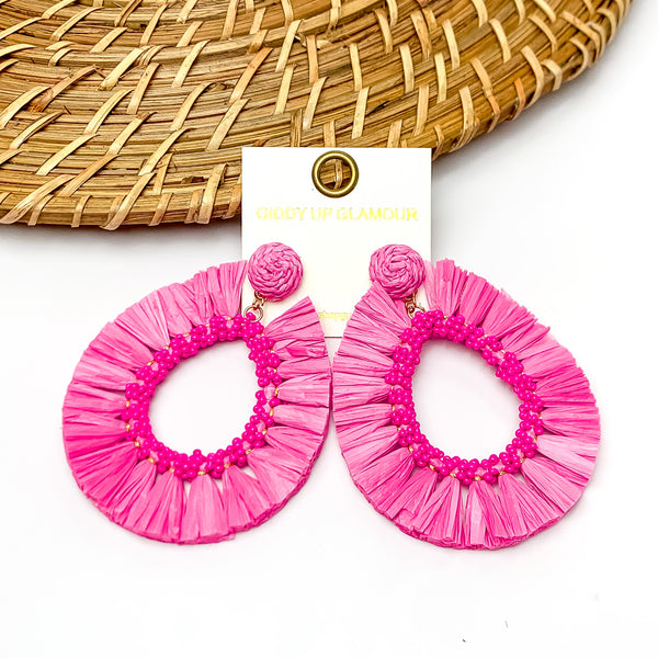 Siesta Sunsets Raffia Wrapped Open Teardrop Earrings in Fuchsia. Pictured on a white background with a wood like decoration at the top.