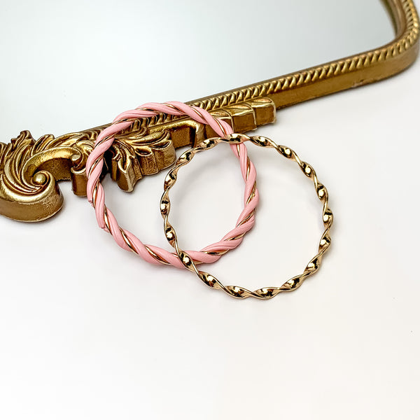 Pictured is a gold twist bangle and gold and pink twist bangle. These bangles are pictured partially leaning on a gold mirror on a white background. 