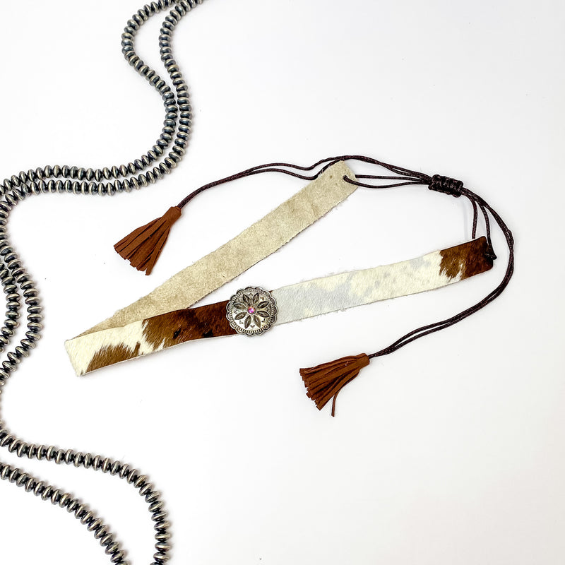 Pictured is a cow print, adjustable hat band with a silver concho charm. This hat band is pictured on a white background with silver, saucer beads on the left side of the hat band. 