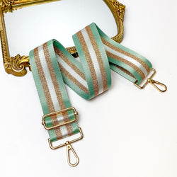 Pictured is a striped purse strap with gold accessories. This purse strap has two outer pink stripes, then two gold glitter stripes, and a center white stripe. This purse strap is laid out partially on a gold mirror on a white background. 