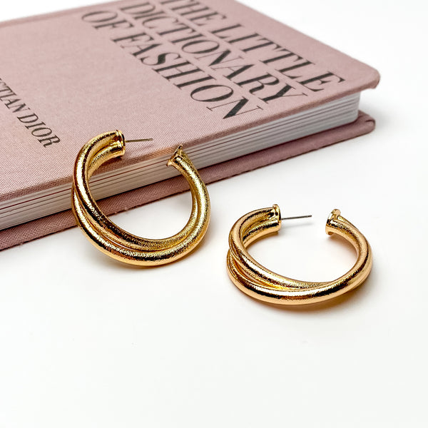 Gold, textured twisted hoop earrings. These earrings are pictured on a white background with one hoop is laying against a mauve colored book. 