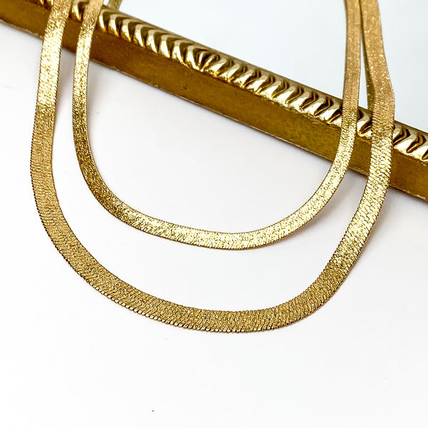 Double layered gold herringbone chain necklace. This necklace is pictured partially laying on a gold mirror on a white background. 