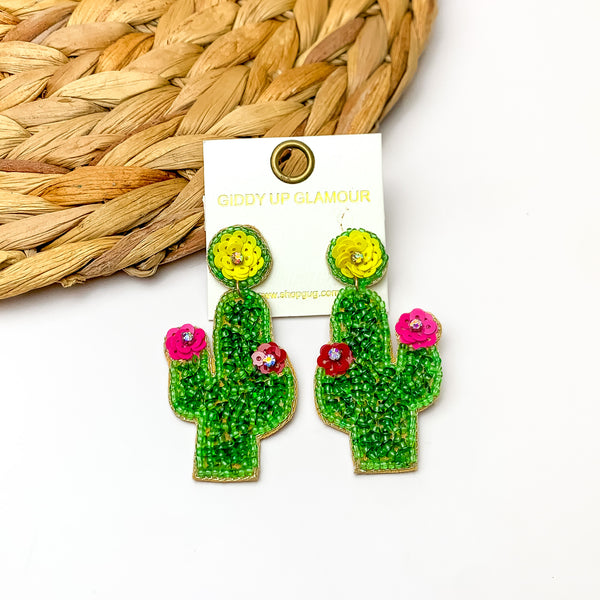 Beaded Cactus Earrings With Multicolor Flowers in Green. Pictured on a white background with a wood like decoration in the top left corner. 
