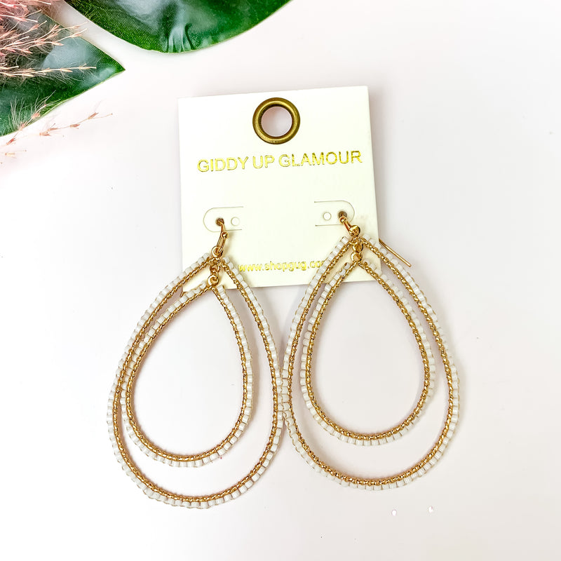Double Open Teardrop Gold Tone Earrings with Beaded Outline in White. Pictured on a white background with leaves in the top left.