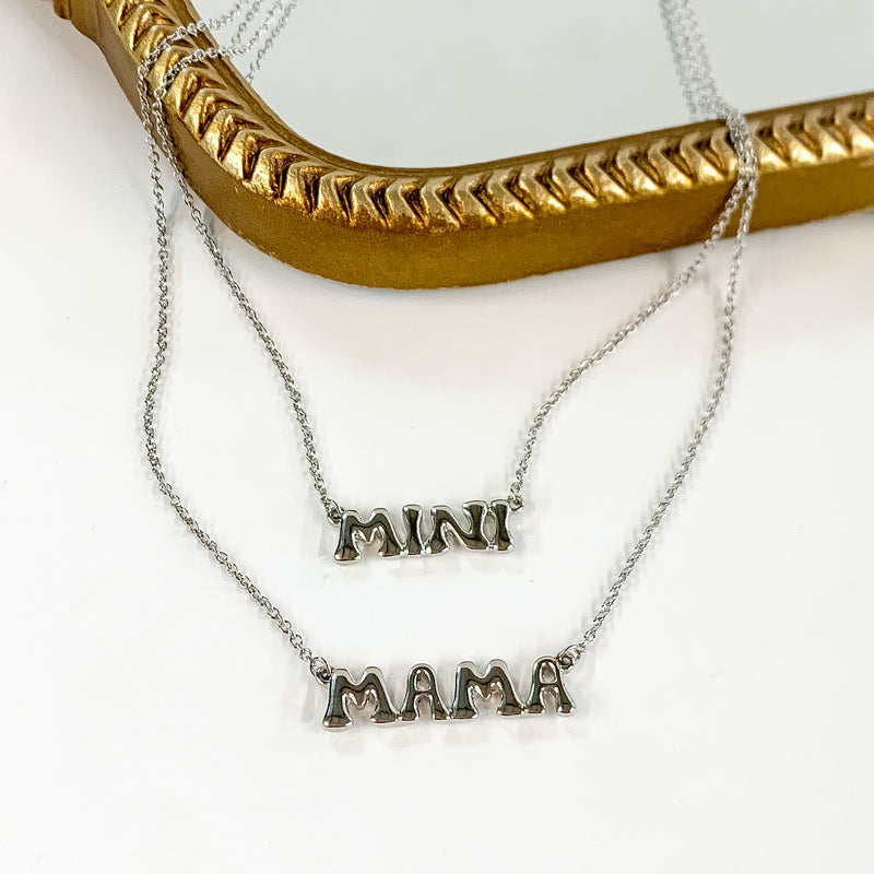 Two silver chain necklaces with word plates. The longer necklace says "MAMA" and the shorter necklace says "MINI." These neckalces are pictured partially laying on a gold mirror on a white background. 