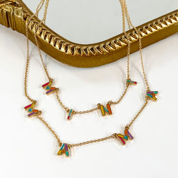 Two gold chain necklaces with multicolored letters. The longer necklace spells out "MAMA" and the shorter necklace says "MINI." These neckalces are pictured partially laying on a gold mirror on a white background. 