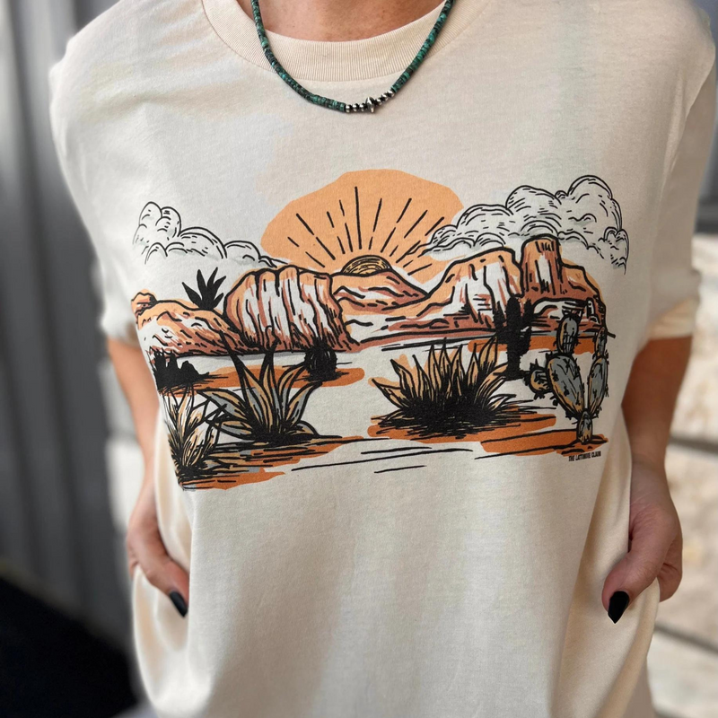 A cream colored short sleeve crewneck tee with a graphic in the center featuring cacti, desert flora, cliffs, and a sunset. Item is pictured on a multicolor background with white, grey, and black