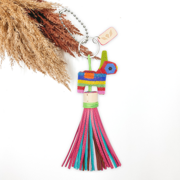 Silver key ring with a beaded donkey charm that has a multicolor rainbow design. There are also blue and pink tassels hanging under the donkey charm. This charm is pictured in front of pompous grass on a white background.