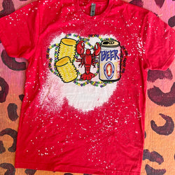 A red short sleeve tee with a bleach splatter pattern and a large bleach spot in the center featuring a graphic of a red crawfish with two corn on the cobs and a beer. Behind the crawfish are purple, green, and yellow mardi gras beads