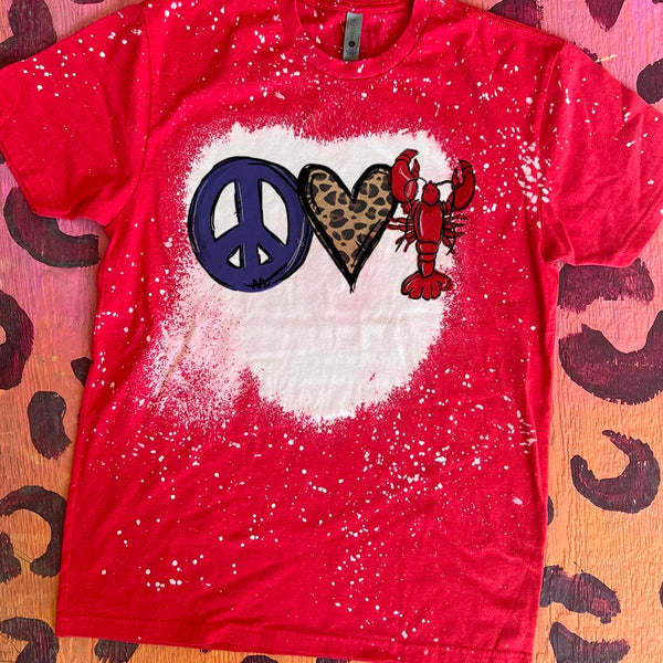 A red short sleeve tee featuring a bleach splatter design with a large bleached spot in the center. In the middle of the bleach spot is a purple peace sign, a cheetah print heart, and a red lobster.