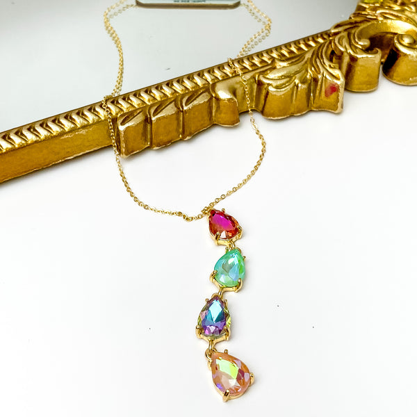 Gold chain necklace with a four crystal drop. This drop are teardrop crystals and include the colors pink, mint, purple, and light pink. This necklace is pictured partially laying on a gold mirror on a white background. 