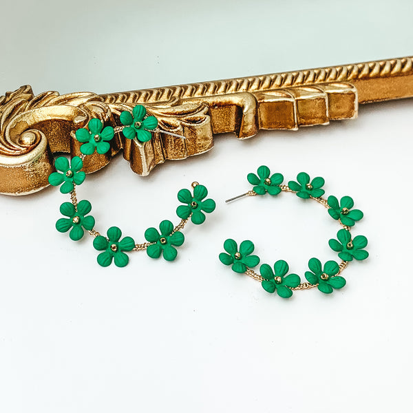 Gold hoop earrings with kelly green flower charms outlining the earrings. These flowers have a gold bead center. One hoop is pictured laying against a gold mirror while the other hoop is laying flat on the white background. 