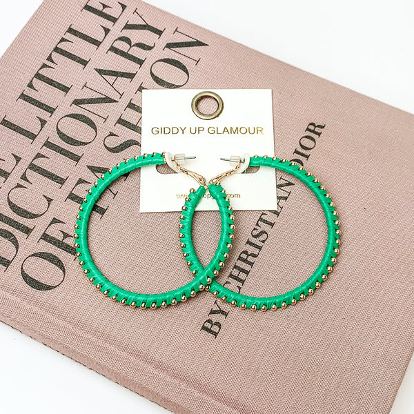 Pictured are circle green hoop earrings with gold beades around it. They are pictured with a pink fashion journal on a white background.