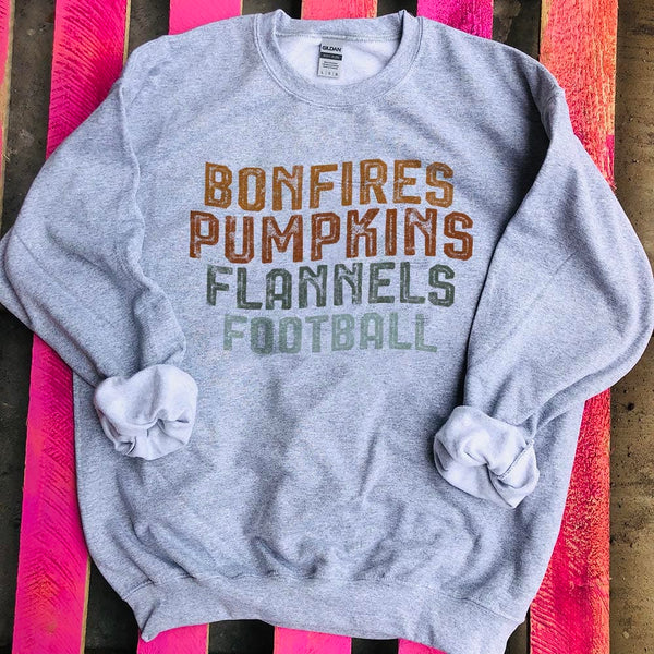 This grey sweatshirt includes a crew neckline, long sleeves, and cute hand drawn design of the following words stacked on top of each other, "Bonfires Pumpkins Flannels, Football" in fun, Fall colors. This is shown here as a flat lay with rolled sleeves. 