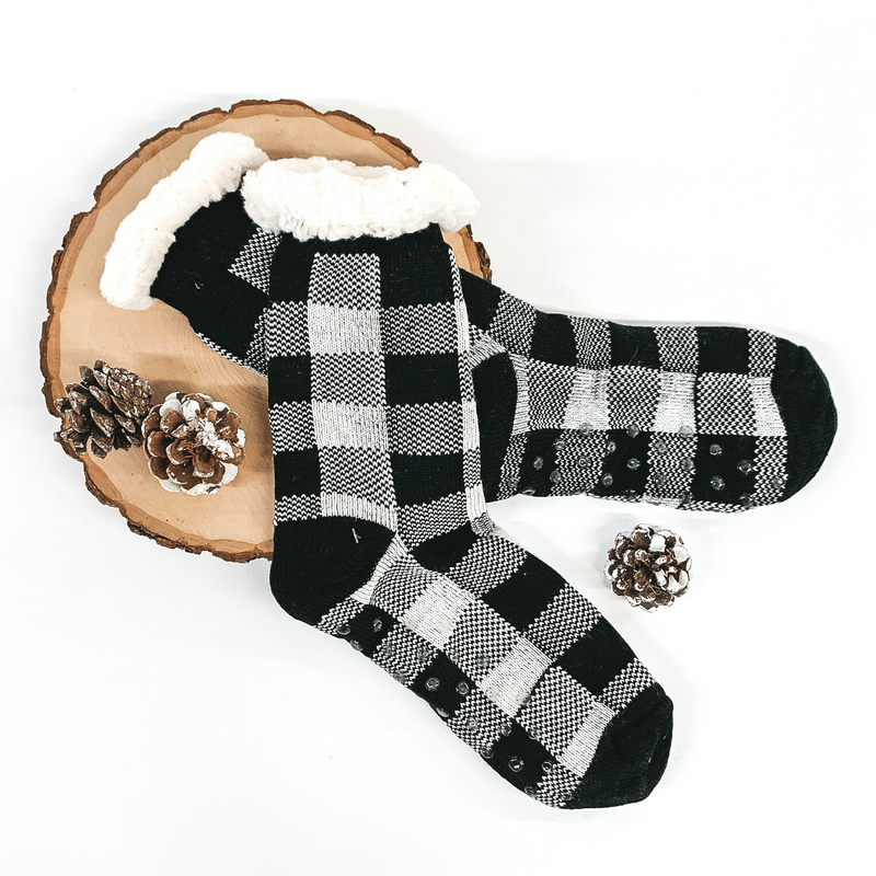 Black and white buffalo plaid socks. The socks has white sherpa at the top of the socks. The socks are pictured on a white background laying on a piece of wood with a few pine cones.