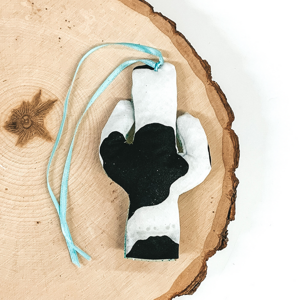 Cactus shaped freshie that is a black and white cow print with light blue string. This is laying on a piece of wood that is on a white background.