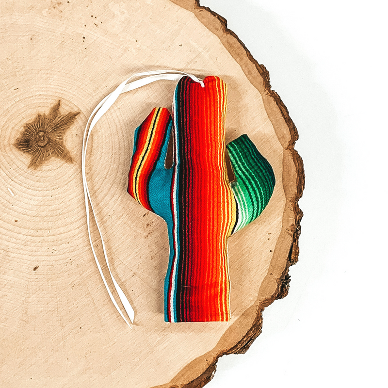Cactus shaped freshie that is a serape print with light pink string. This is laying on a piece of wood that is on a white background.